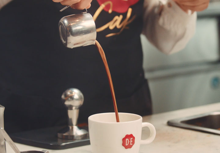 Persoon met Douwe Egberts schort die koffie toevoegt aan een koffiemok